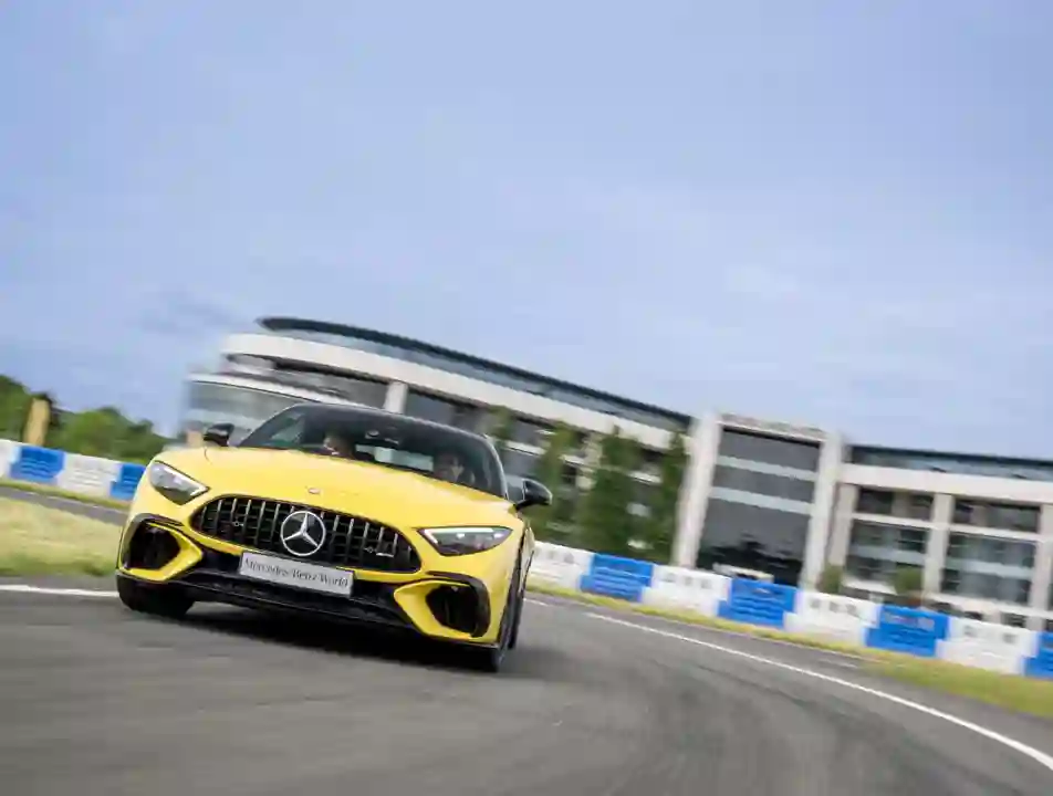 Bright yellow Mercedes-AMG speeding around a bend at Mercedes-Benz World, with driver and passenger enjoying a dynamic track experience in front of a modern building backdrop.