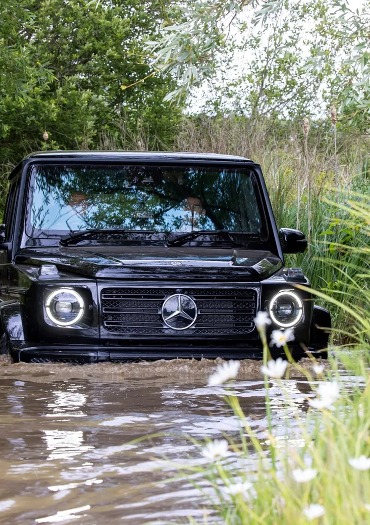Black Mercedes-Benz G-Class SUV driving through deep muddy water during an off-road experience, surrounded by dense greenery and wildflowers.