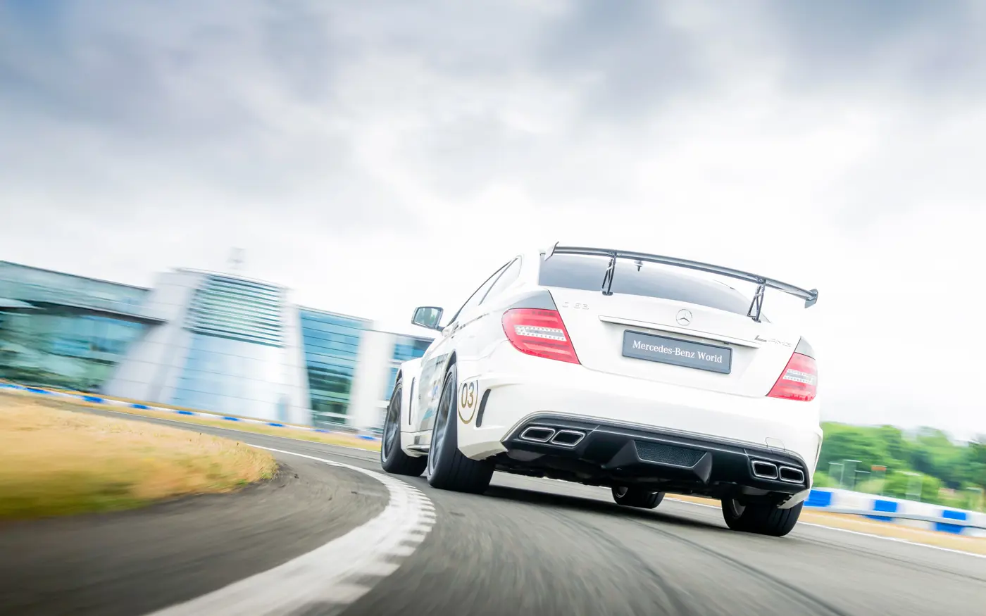 Rear view of a white Mercedes-AMG C63 with racing livery and rear spoiler, taking a sharp corner on the track at Mercedes-Benz World with motion blur.