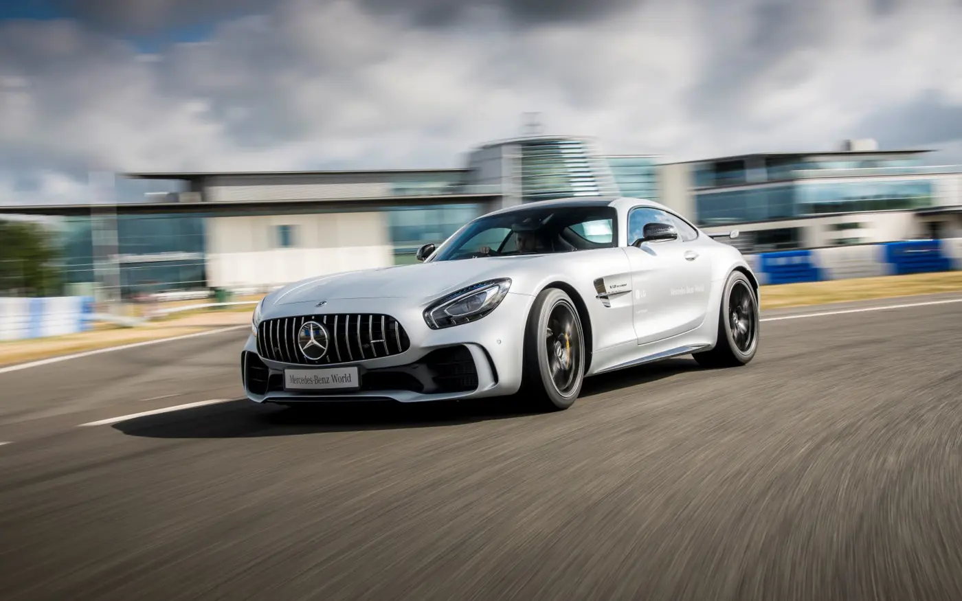 Silver Mercedes-AMG GT speeding around a track at Mercedes-Benz World, with motion blur highlighting its performance and sleek design against a modern building backdrop.