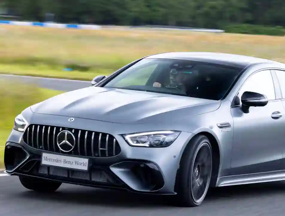 A silver Mercedes-AMG speeding on a test track at Mercedes-Benz World, with the driver focused and blurred trees and grass in the background.