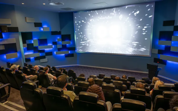 Audience seated in a modern cinema with blue accent lighting, watching a film on a large screen in a comfortable, dimly lit theatre setting.