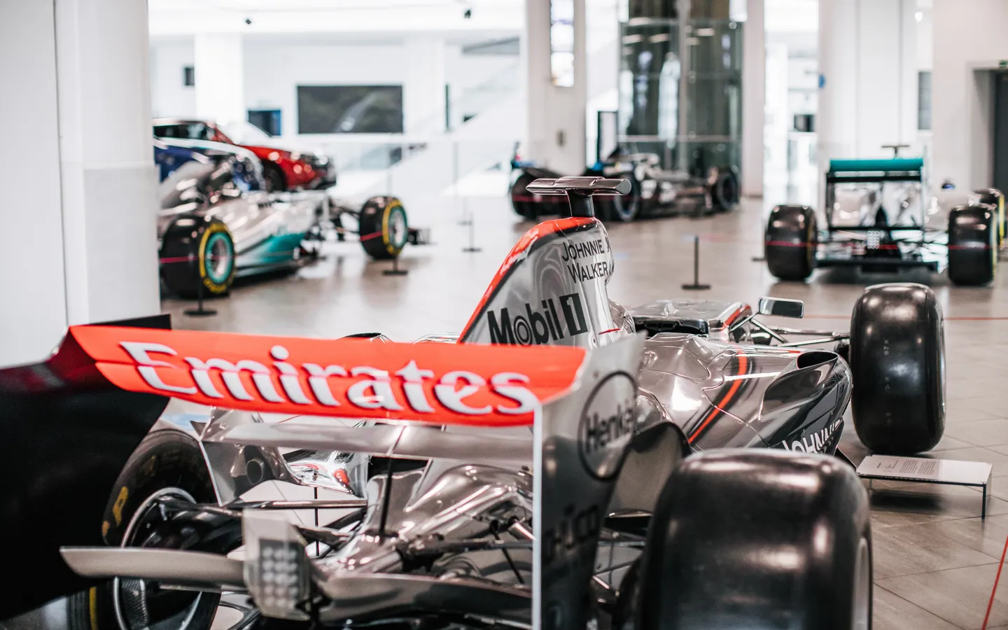 A close-up view of a silver and red McLaren Mercedes Formula 1 car on display, surrounded by other F1 race cars in a sleek, modern exhibition hall.