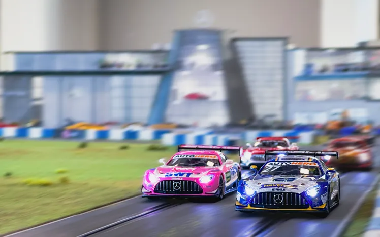 A detailed model race track featuring miniature Mercedes-Benz DTM cars in front of a scale replica of the Mercedes-Benz World building.