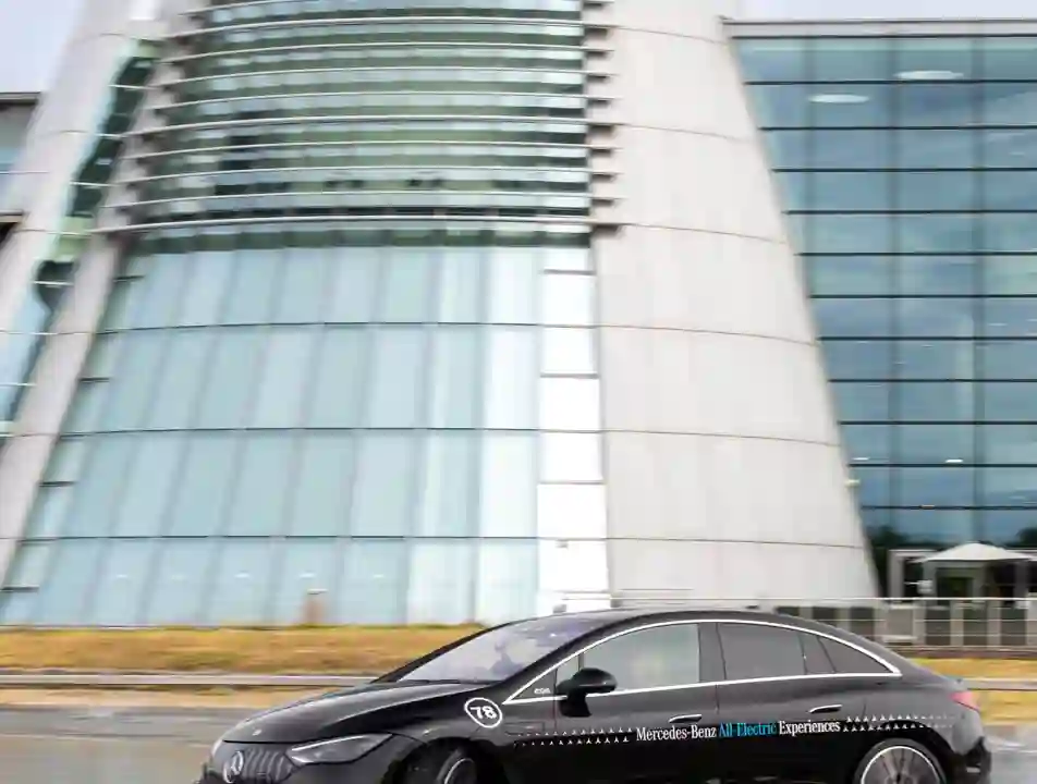 A black Mercedes-Benz EQE electric car drives through water on a test track in front of the iconic glass-fronted Mercedes-Benz World building.