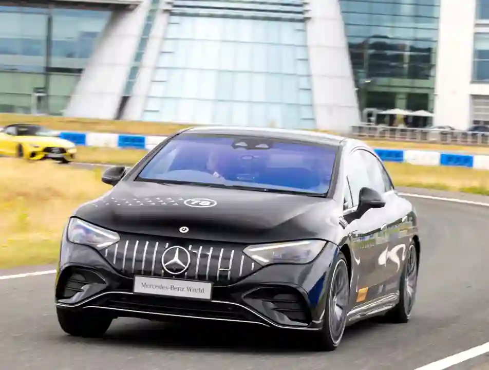 A black Mercedes-Benz car speeds along a track outside Mercedes-Benz World, with the building's iconic glass façade and logo visible in the background.