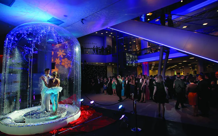 A crowd watches a theatrical performance inside a giant snow globe at a glamorous indoor event, with elegant lighting and guests dressed in formal attire.
