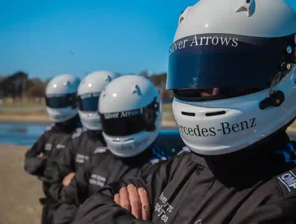 Silver Arrows driving team in full racing gear with helmets and Mercedes-Benz branded suits, standing in formation under a clear blue sky.