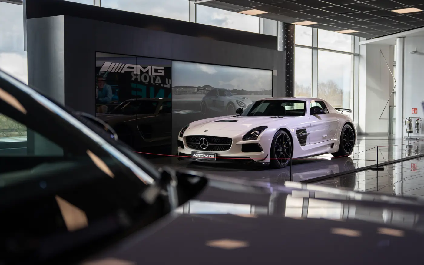 White Mercedes-AMG SLS Black Series on display inside Mercedes-Benz World, spotlighted in a showroom setting with reflective floors and digital backdrop.