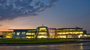 Exterior view of Mercedes-Benz World at dusk, with lights glowing from the glass-fronted building and a large open driving surface in the foreground.