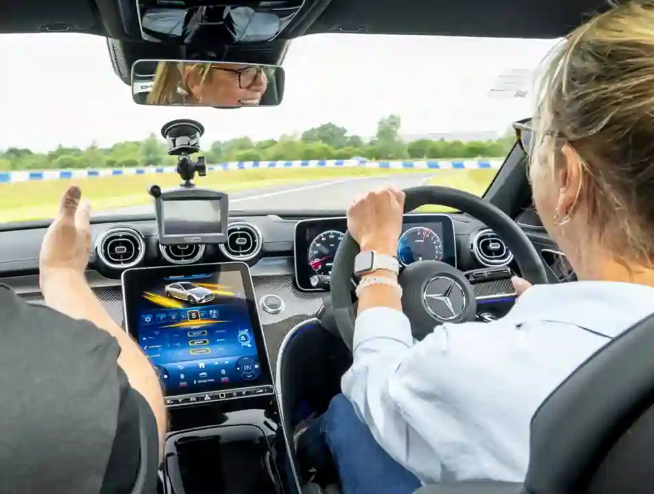 Interior view of a Mercedes-Benz during a driving experience, with a woman at the wheel, instructor beside her, and advanced dashboard and navigation systems visible.