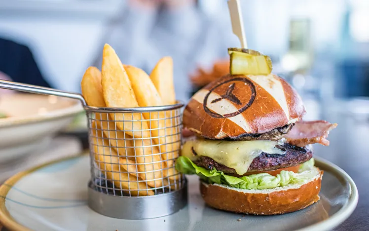 Close-up of gourmet burgers and fries served on a wooden tray in a casual dining setting, with a hand reaching for a drink in the background.