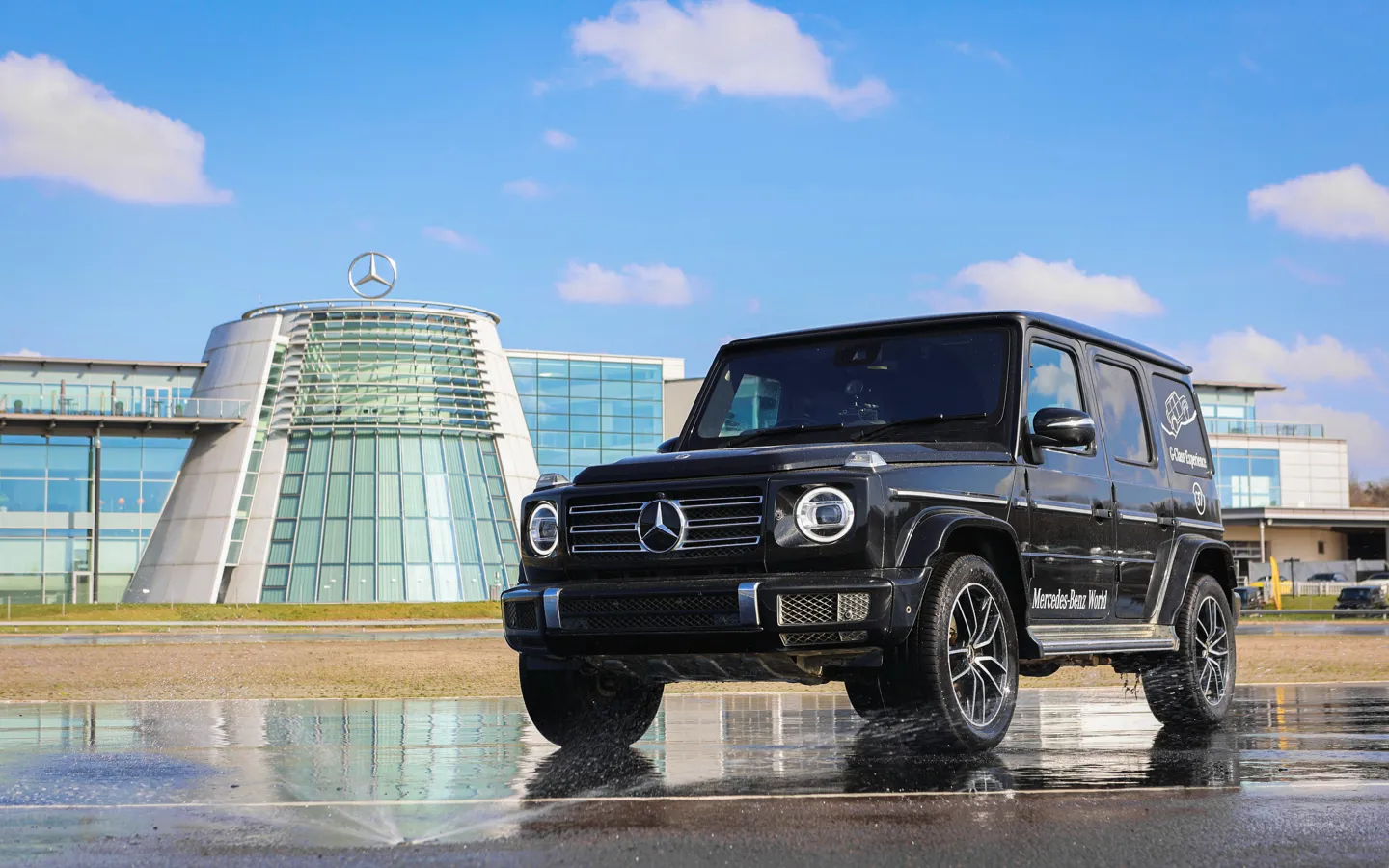 Black Mercedes-Benz G-Class SUV driving through water outside the glass-fronted Mercedes-Benz World building on a sunny day with blue skies.