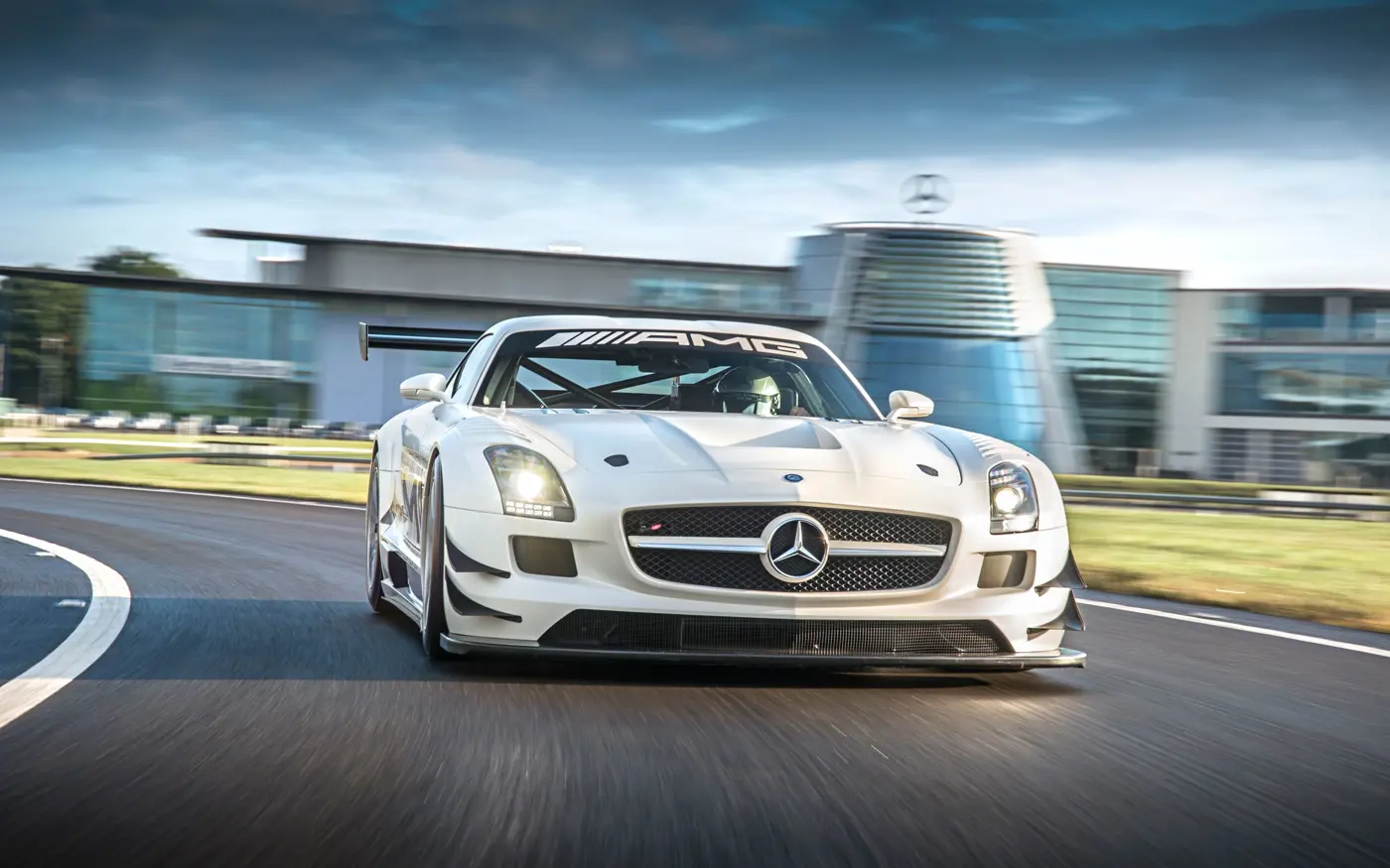 White Mercedes-AMG race car speeding around a track in front of the Mercedes-Benz World building, with a driver in full gear behind the wheel.